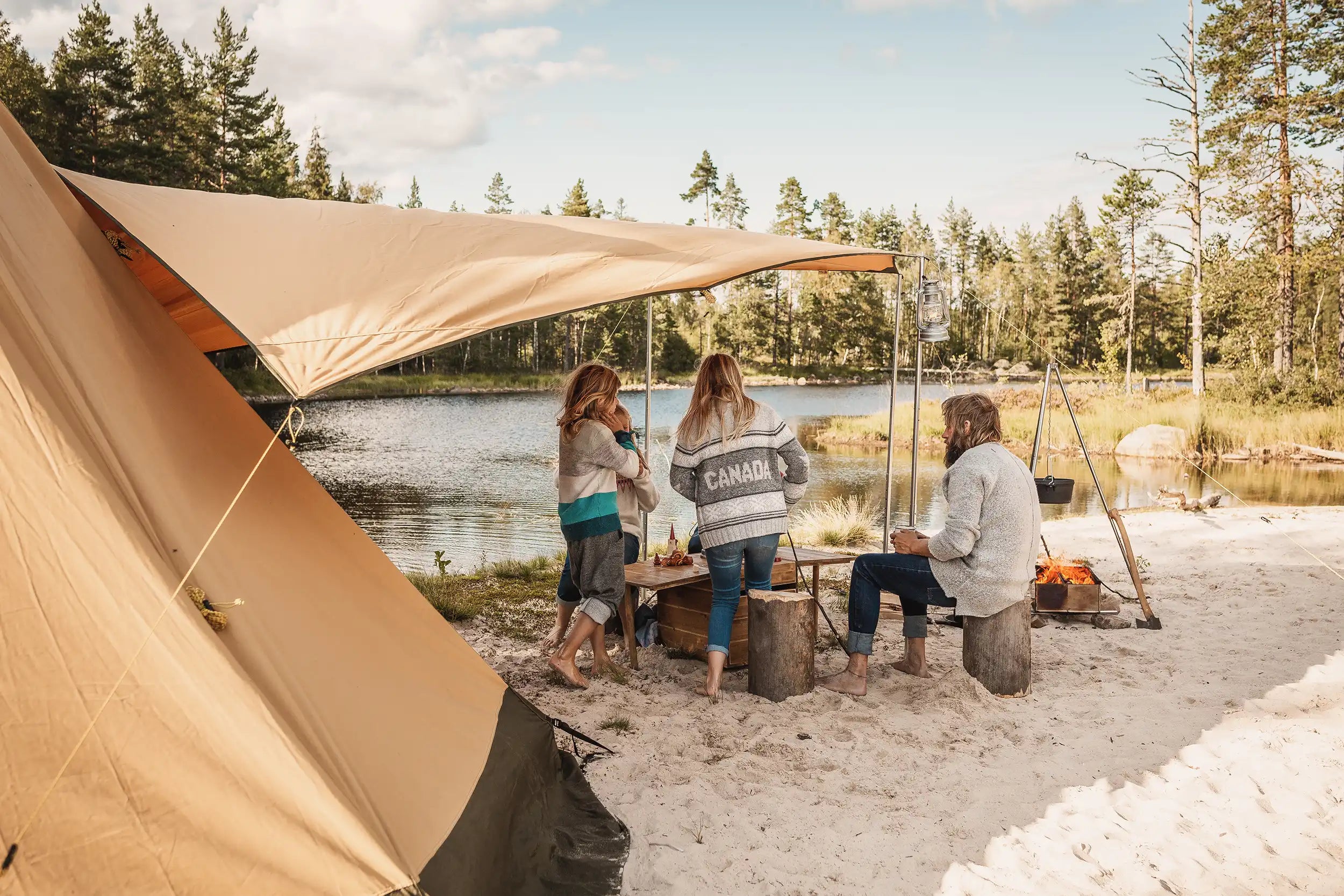 Family camping by a lake with canvas tent pitched on the beach with open fire outside the tent and wood burning stove inside